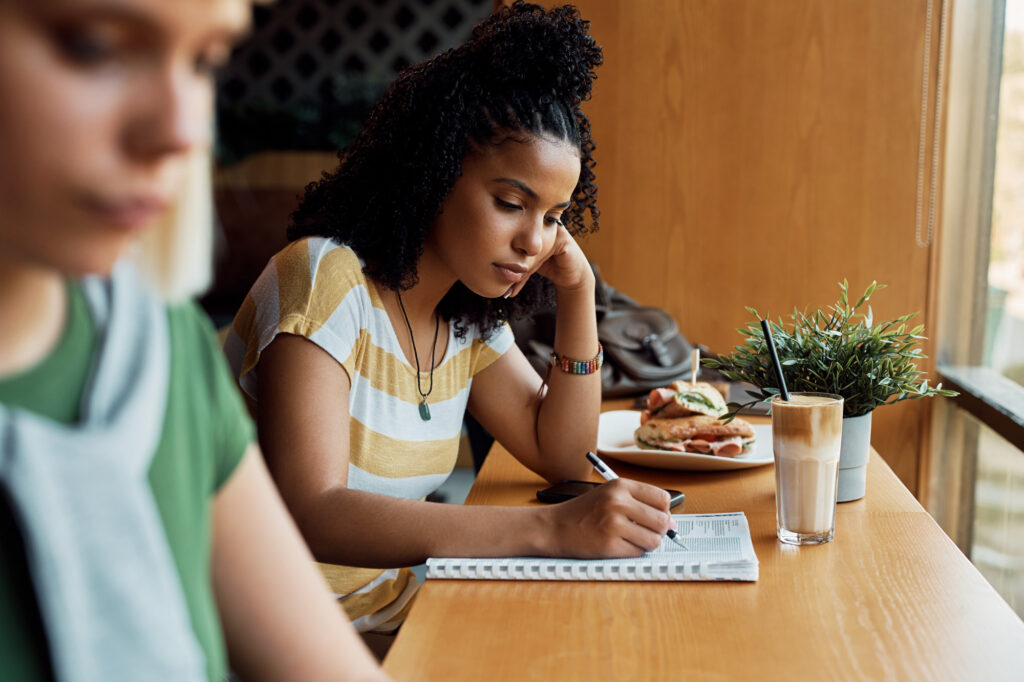 A young woman with curly hair writes in a notebook at a cafe table