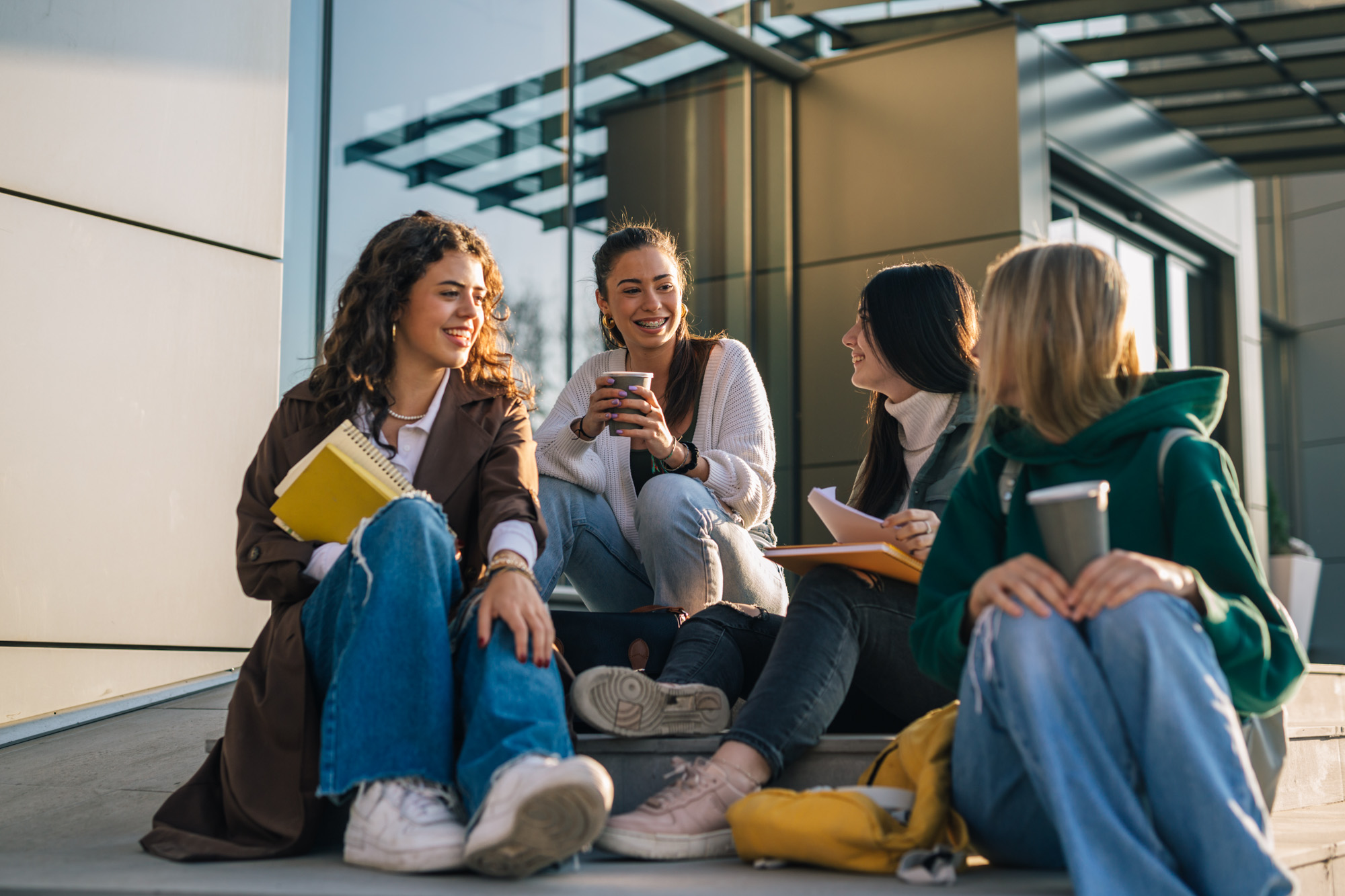 Four female students sitting on outdoor steps talking and drinking coffee together