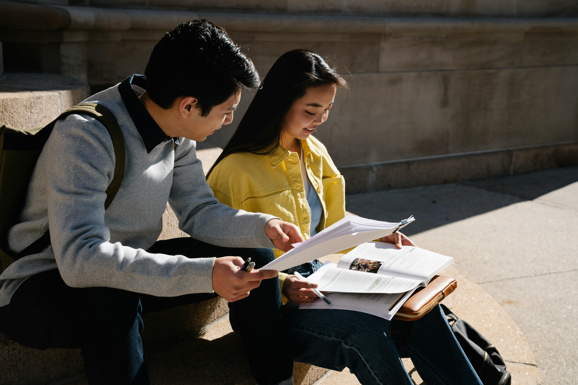 Two students sitting on outdoor steps reviewing paperwork and a textbook together