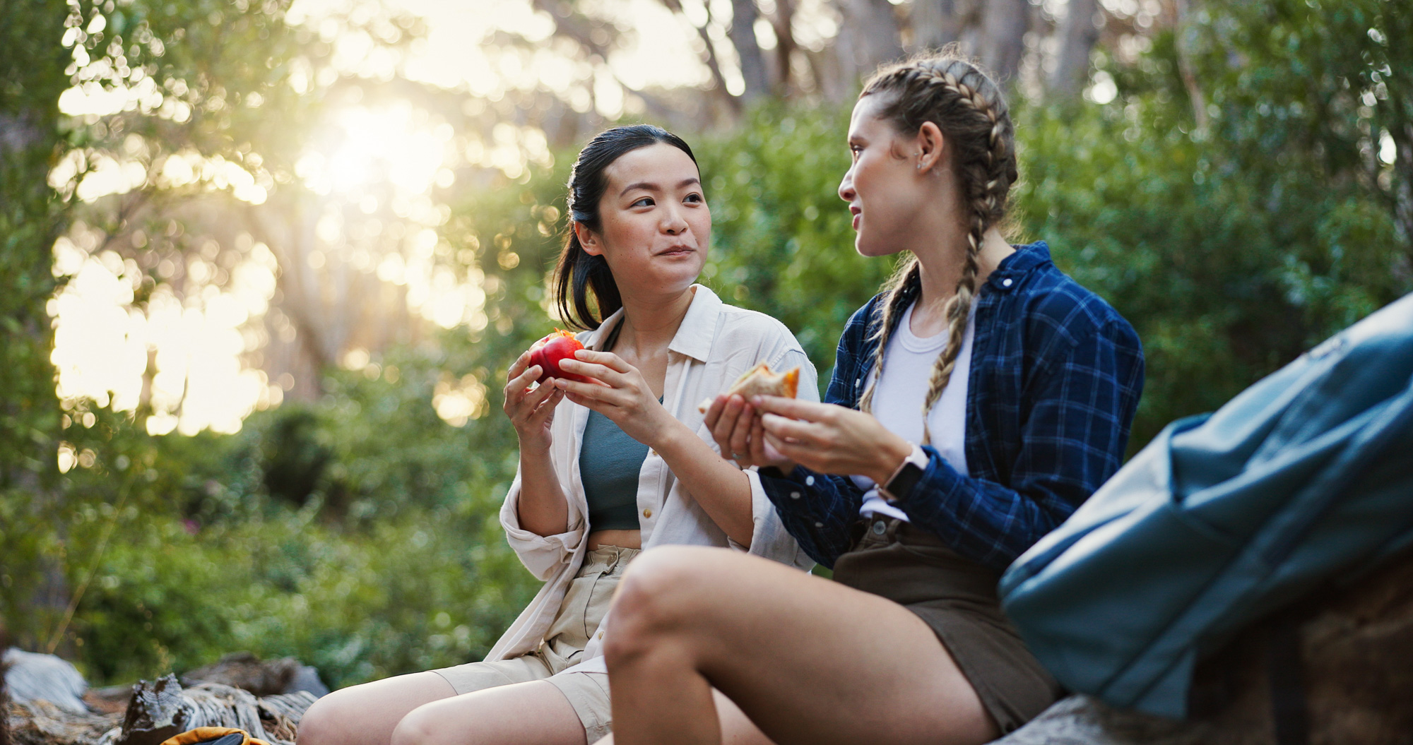 Two women sitting in the woods eating snacks and talking together during a hike
